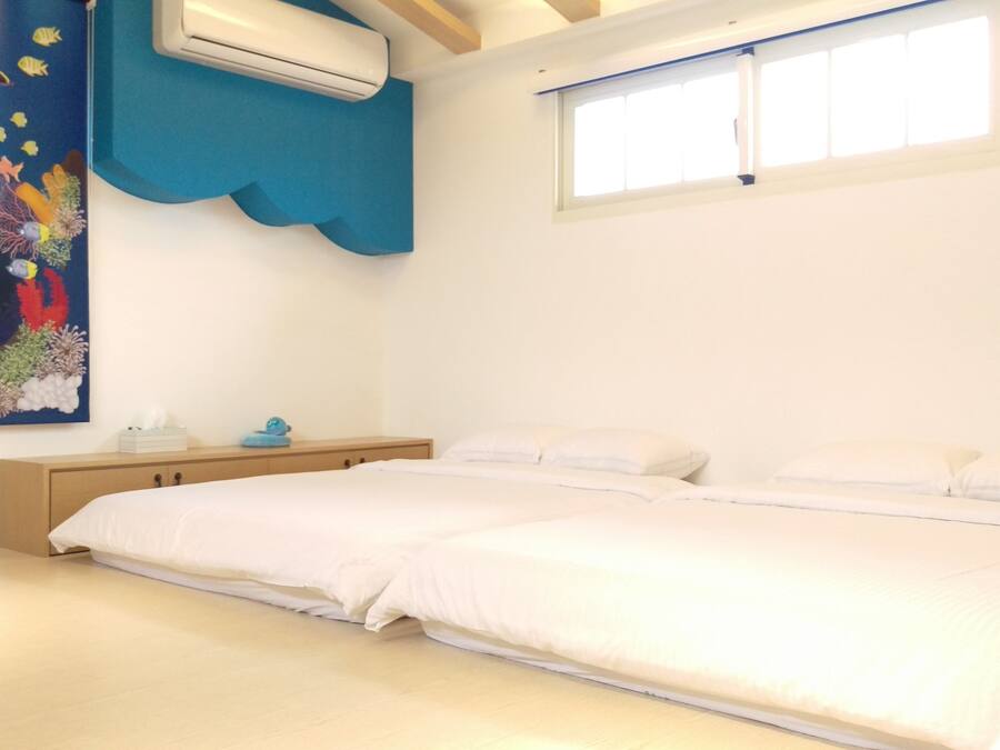 A bedroom with a bed, a nightstand, a wall-mounted air conditioner, a window, and a colorful painting of marine life.