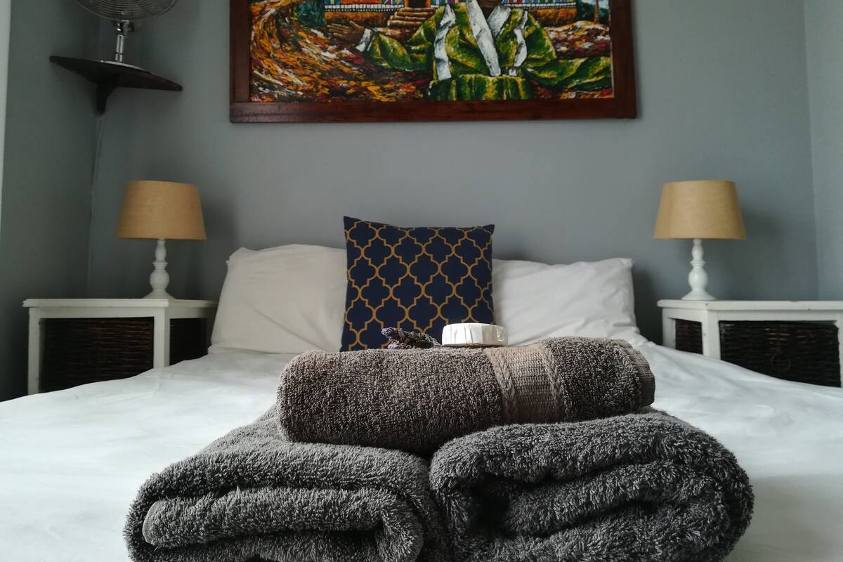 A neatly made bed with two pillows, a wall-mounted fan, and a painting above it.