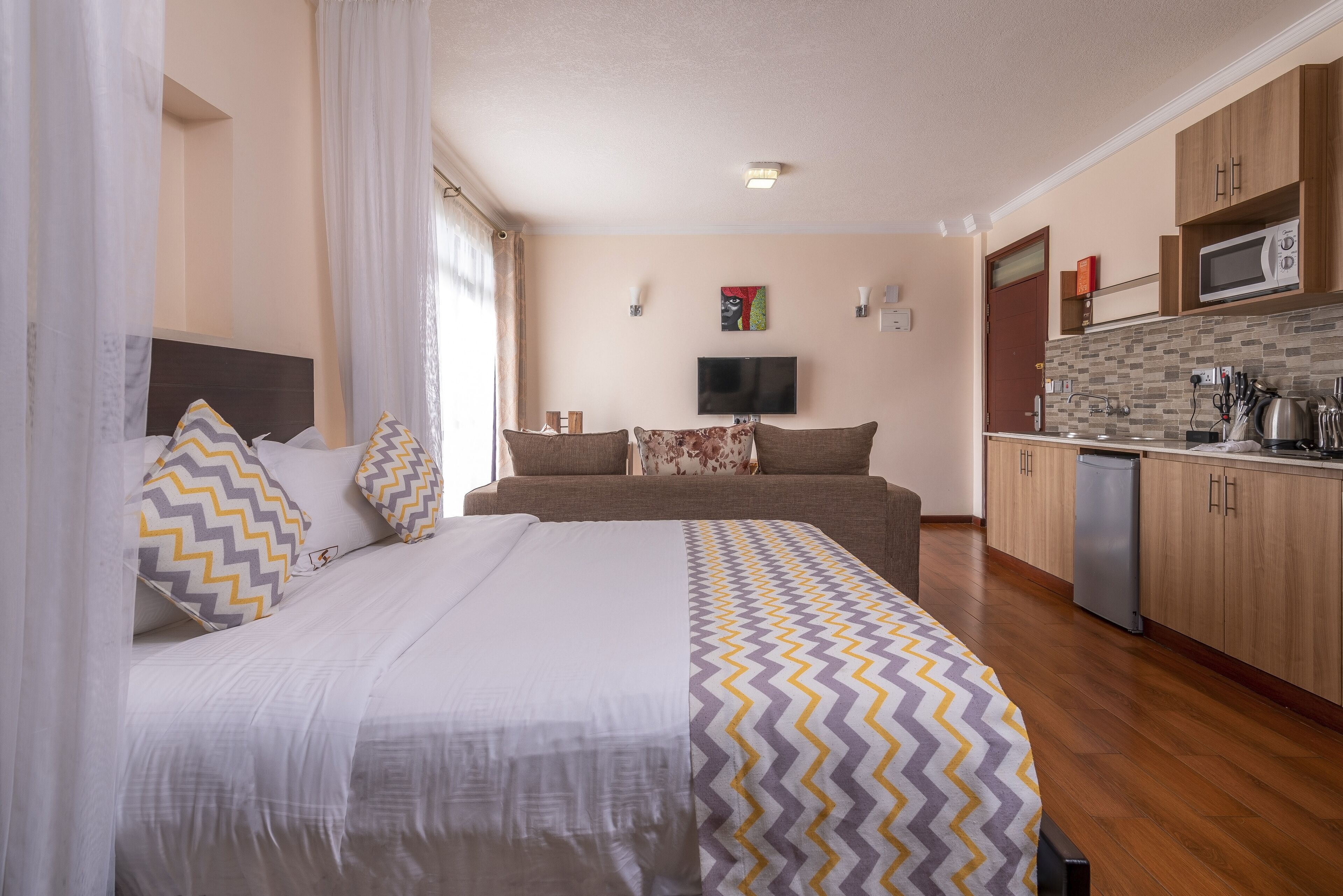 A bedroom with a bed, a sofa, a TV, and a kitchen area with a microwave and sink.