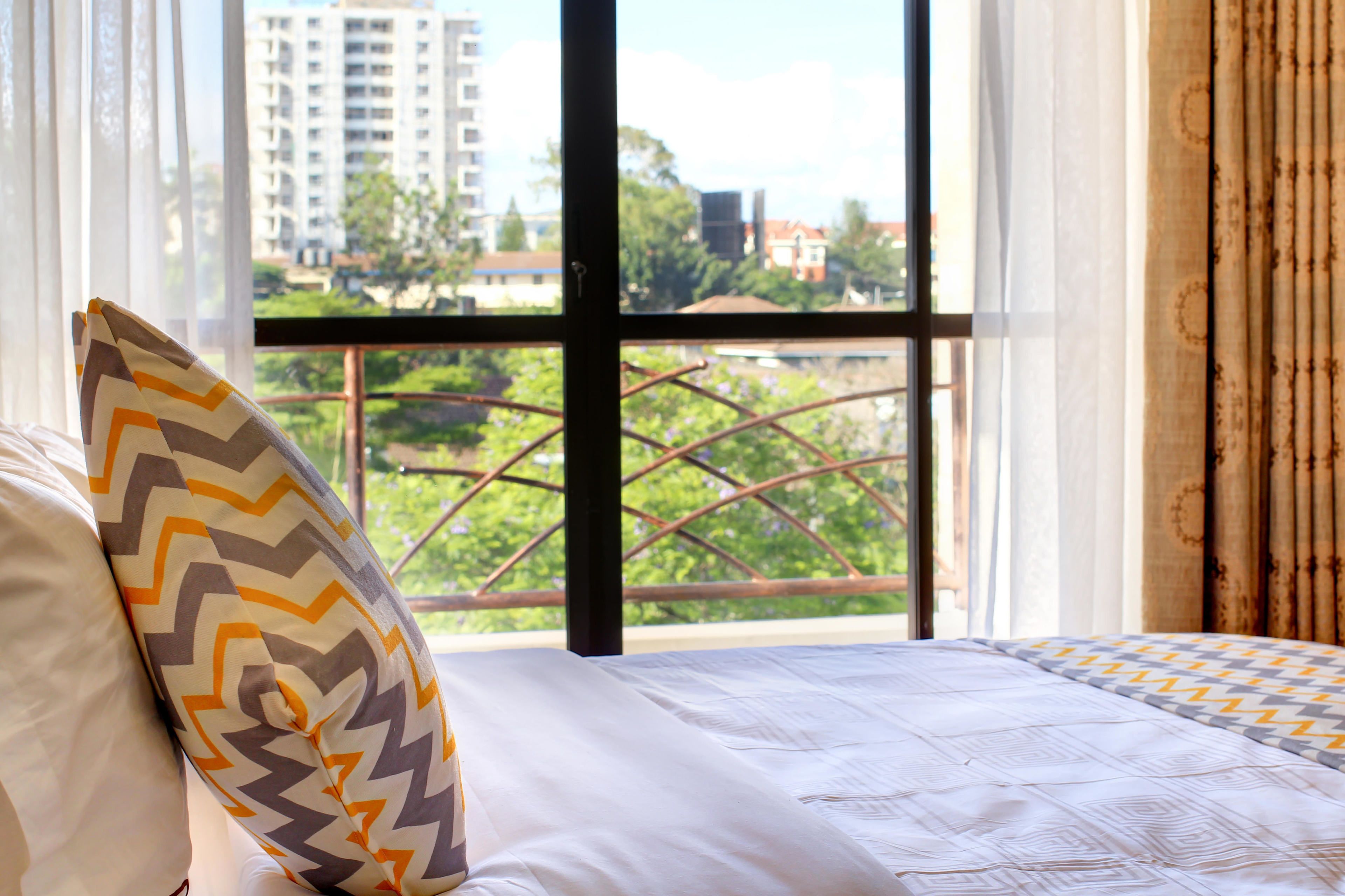 A hotel room with a large window, a bed with patterned bedding, and a view of buildings and greenery outside.