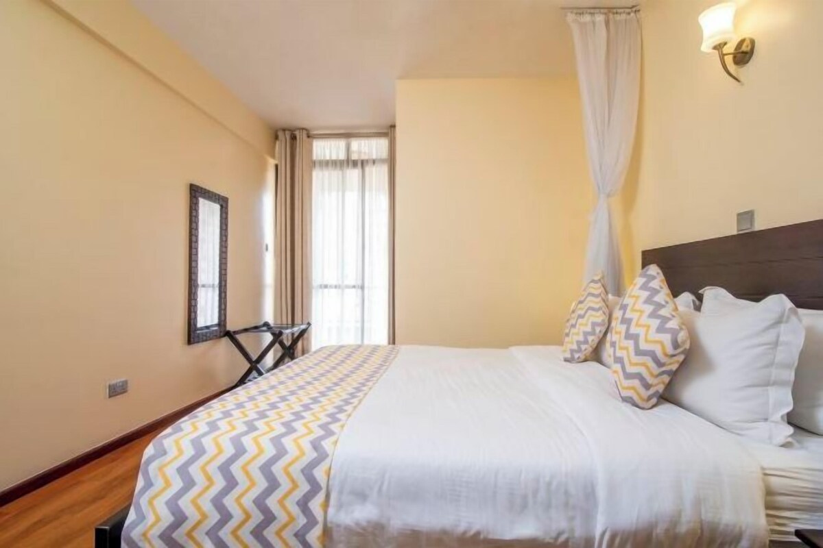 Un dormitorio con cama, mesita de noche, lámpara de pared, ventana con cortinas y espejo.