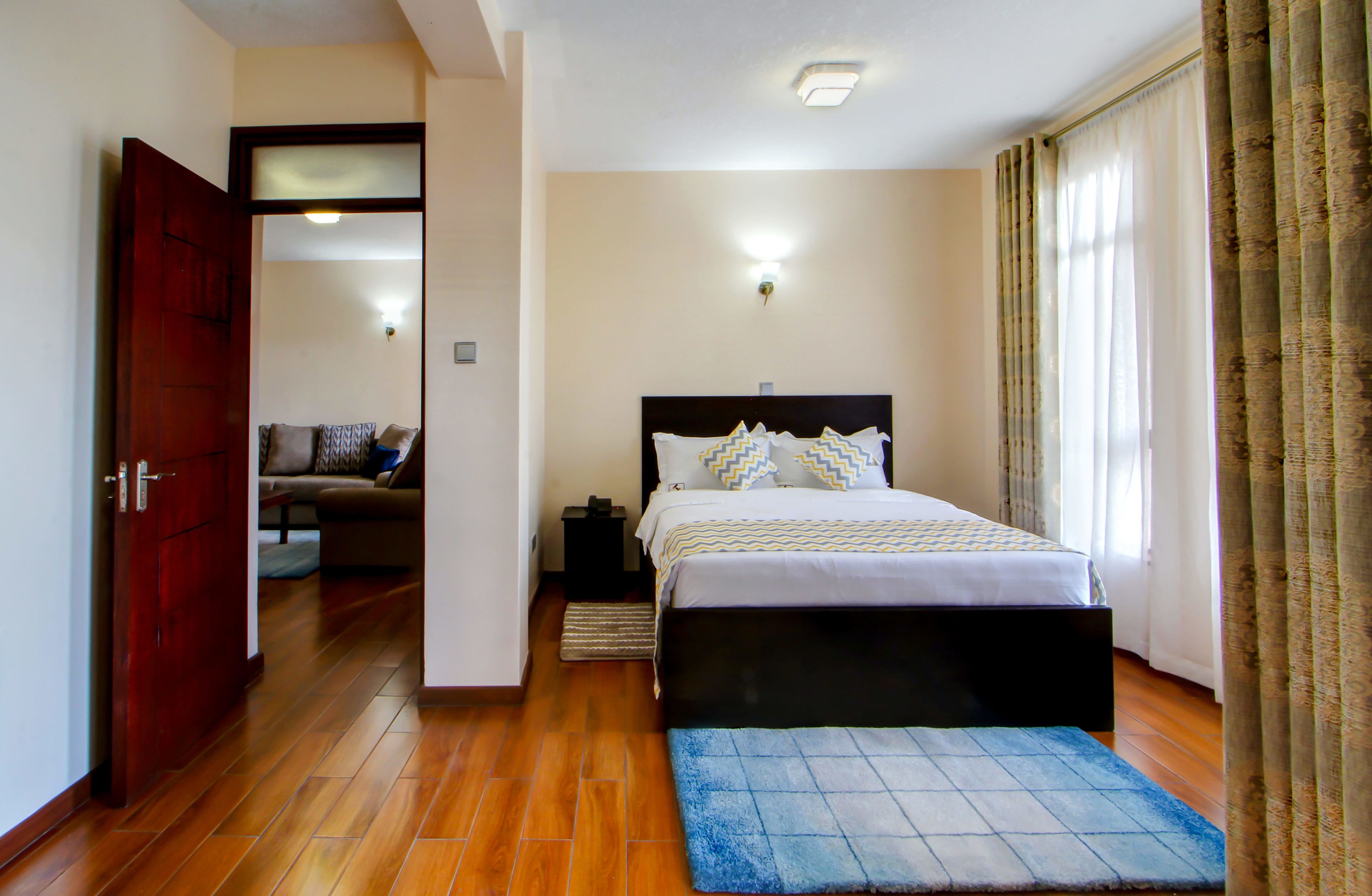 A bedroom with a bed, a nightstand, a wooden floor, a door, and a view of another room.