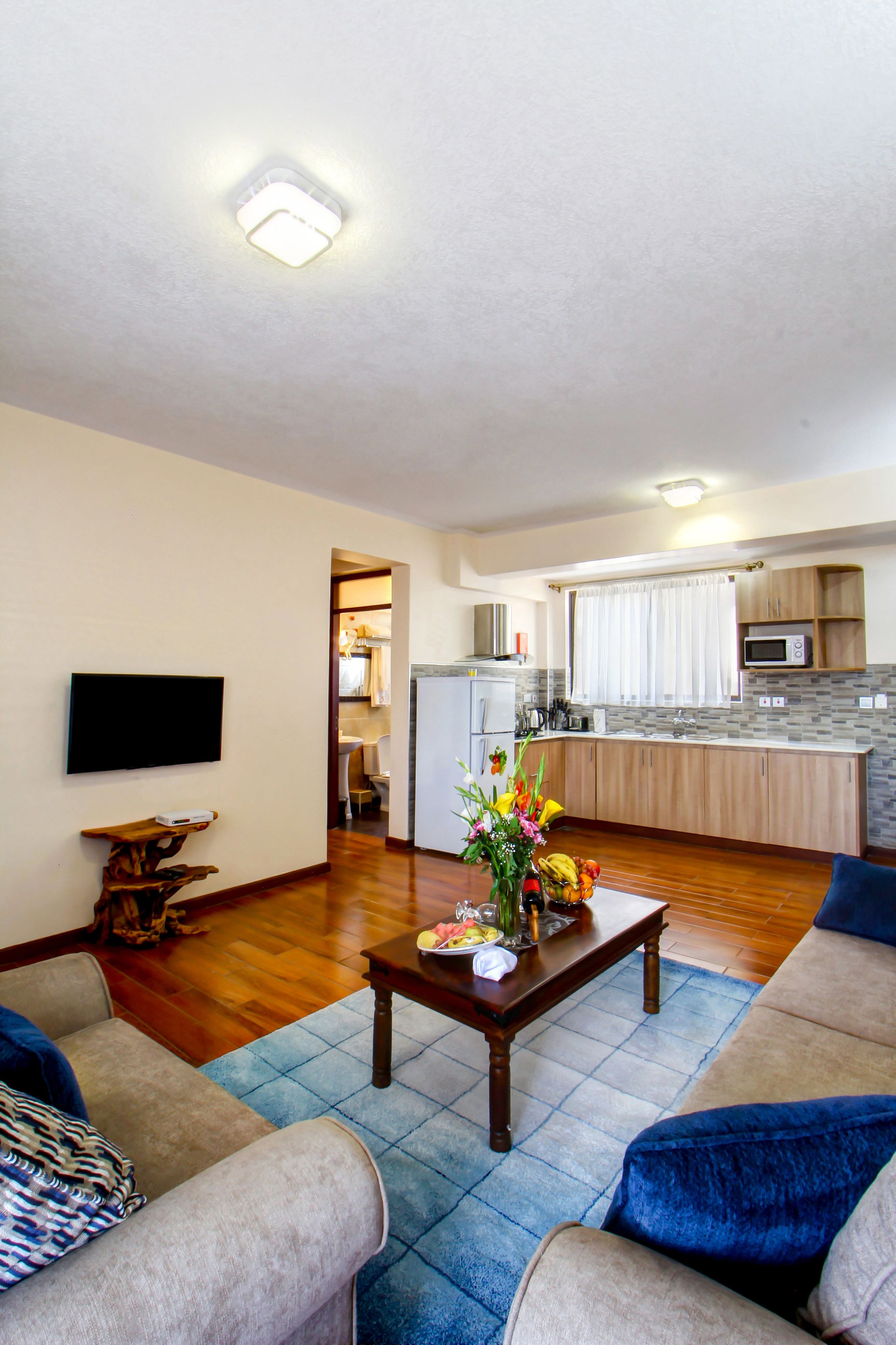 A living room with a sofa, a coffee table with a vase, and a TV on the wall. There is a kitchen in the background with a microwave and cabinets.