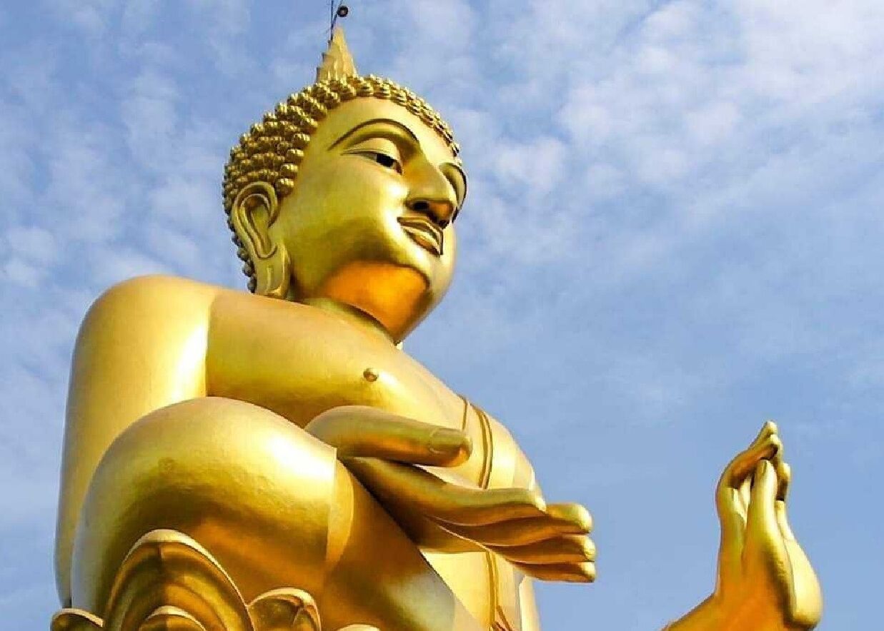 Een groot gouden beeld van een zittende figuur met handen in gebed, voor een heldere lucht.