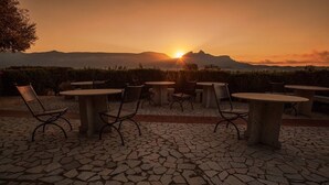 Mobiliario exterior con mesas y sillas, suelo de baldosas de piedra y vistas al atardecer sobre un paisaje montañoso.