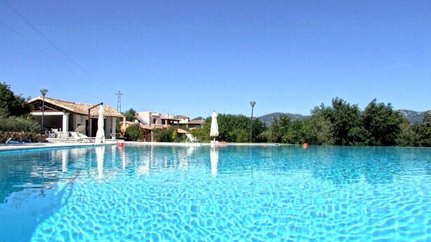 Una piscina azul clara con sillas de descanso y sombrillas, rodeada de edificios y árboles.