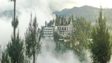 Um edifício com vários andares e design arquitetônico moderno, rodeado por uma floresta densa e montanhas envoltas em névoa.