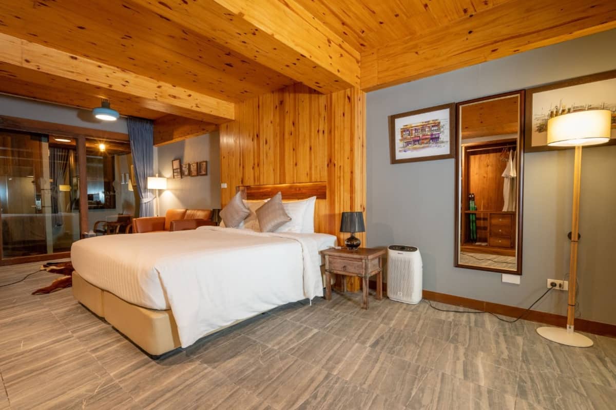 A hotel room with a wooden ceiling, a large bed, a bedside table, a lamp, a mirror, and a view of the outside.