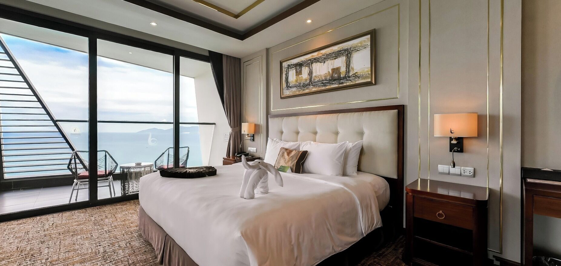 Une chambre d’hôtel avec un grand lit, une vue sur la mer, un coin repas avec une chaise et un tableau au mur.