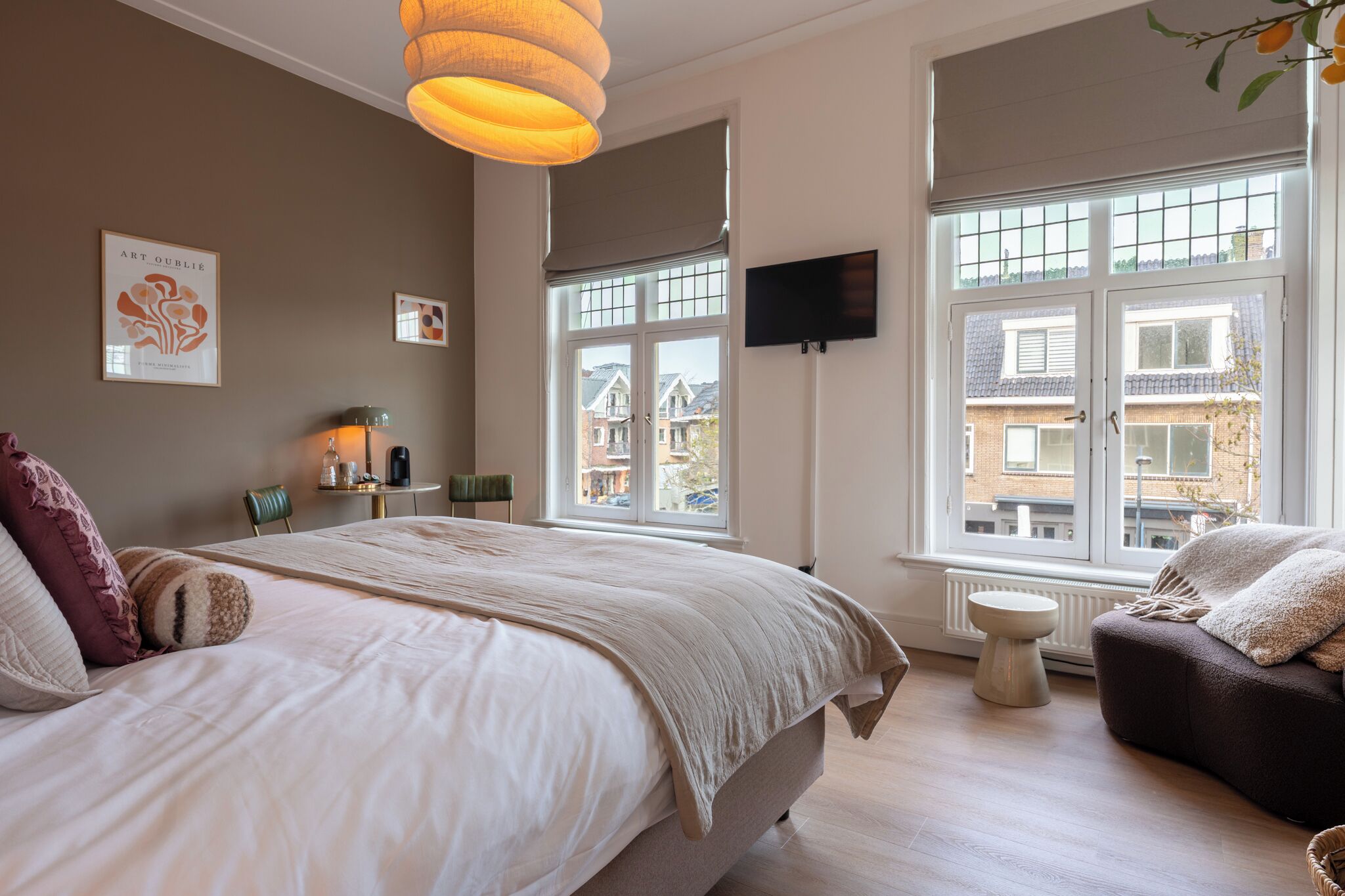 A bedroom with a large bed, a TV mounted on the wall, and a window with a view of buildings outside.