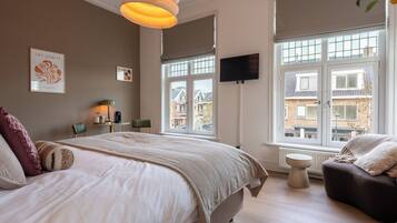 A bedroom with a large bed, a TV mounted on the wall, and a window with a view of buildings outside.