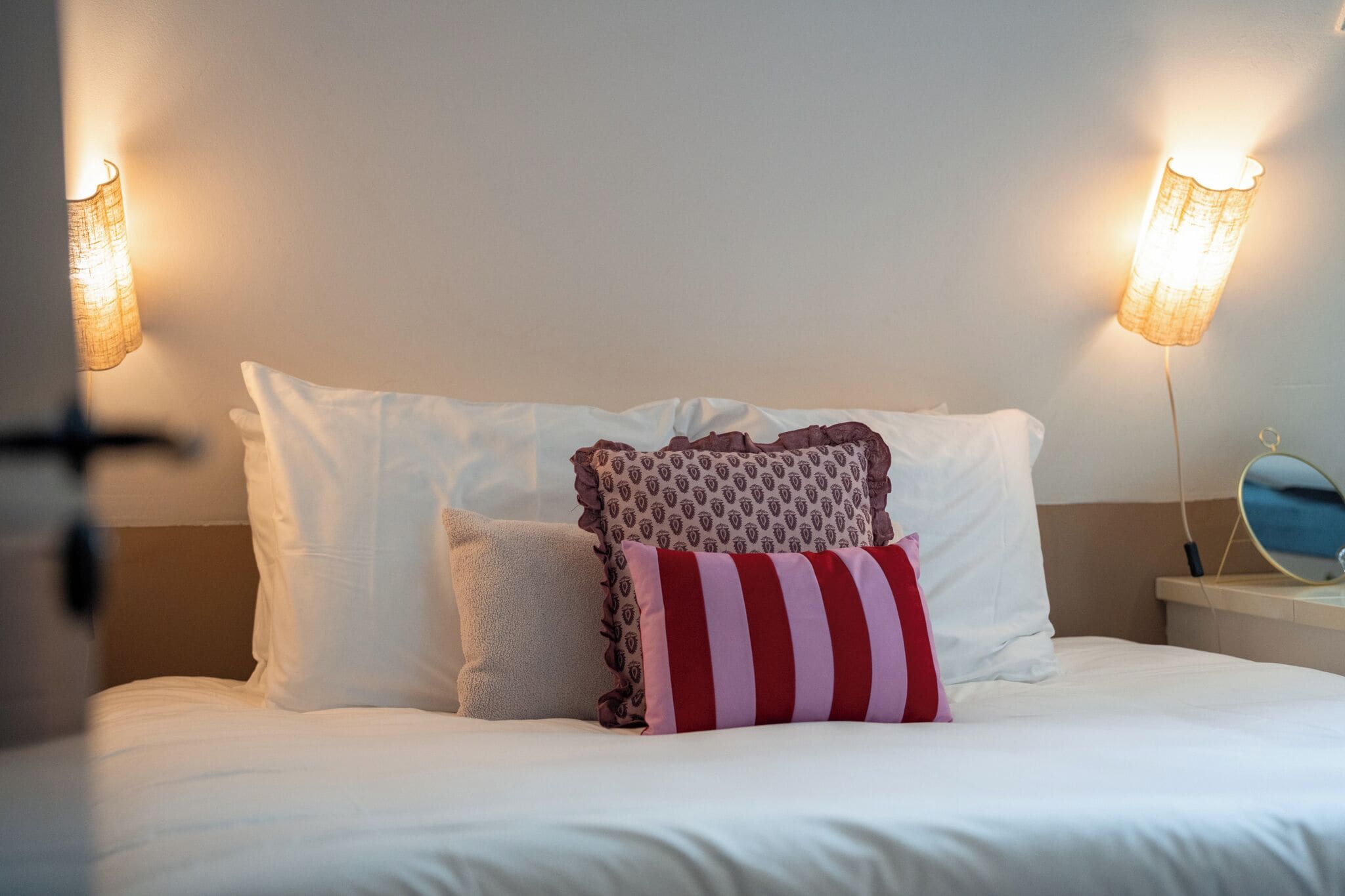 Una cama bien hecha con sábanas blancas y varias almohadas, incluyendo una a rayas. Hay dos lámparas de pared a cada lado de la cama.