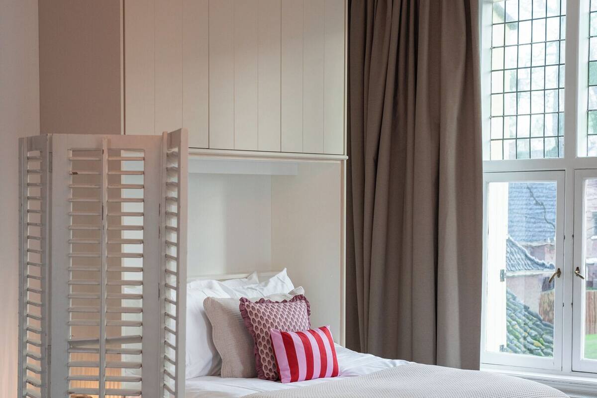 Quarto com cama, guarda-roupa branco, lustre e janela com cortinas.
