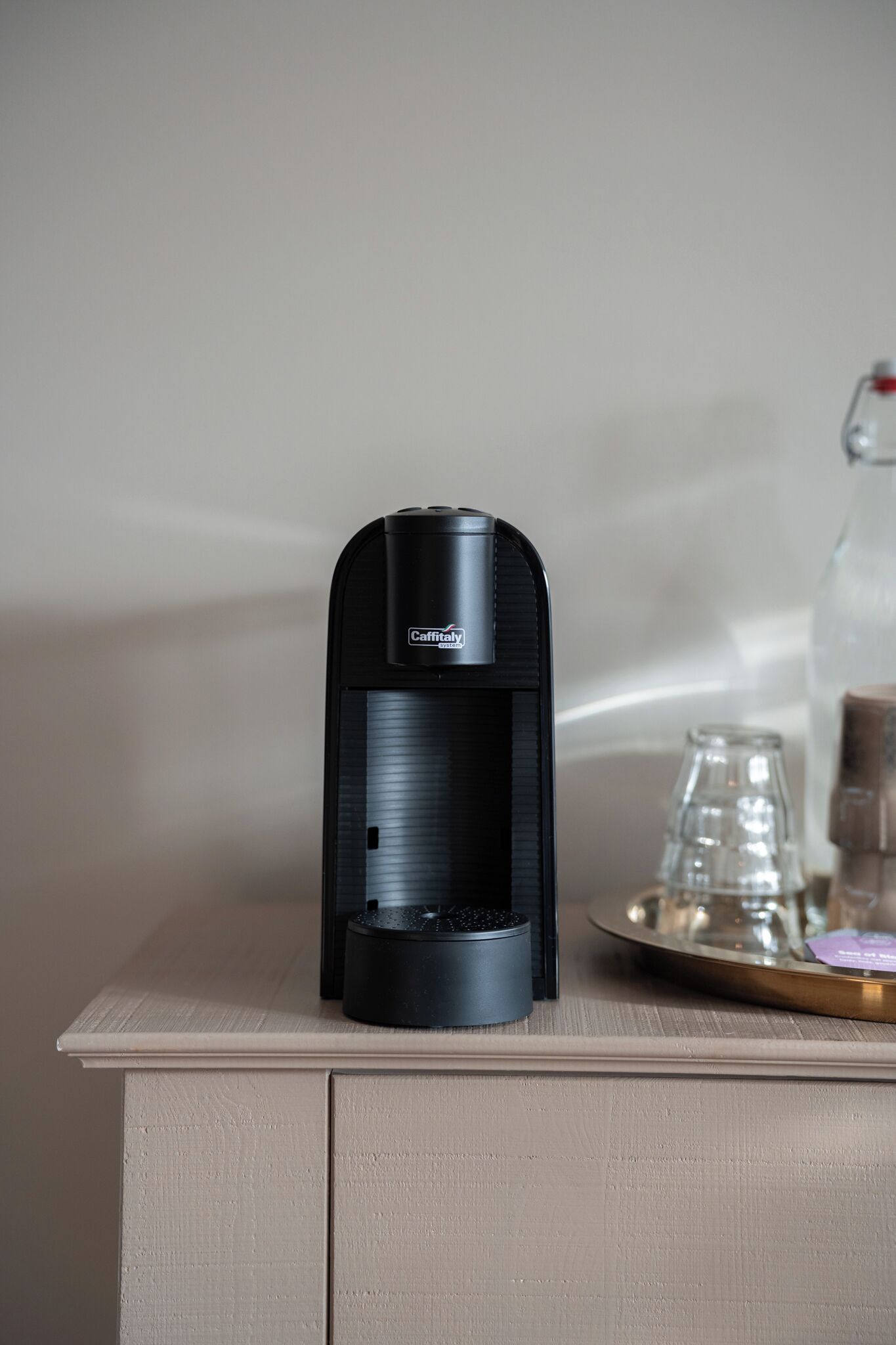 Une machine à café noire est posée sur un meuble de couleur claire. On aperçoit en arrière-plan un verre et une assiette.