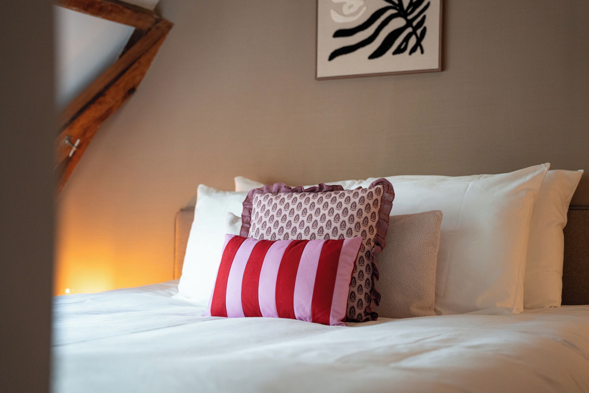 A neatly made bed with white and patterned pillows, a framed artwork on the wall, and a warm light source in the background.