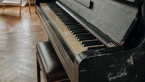 Een vleugelpiano in een kamer met houten vloer, een staande lamp en een raam met gordijnen.