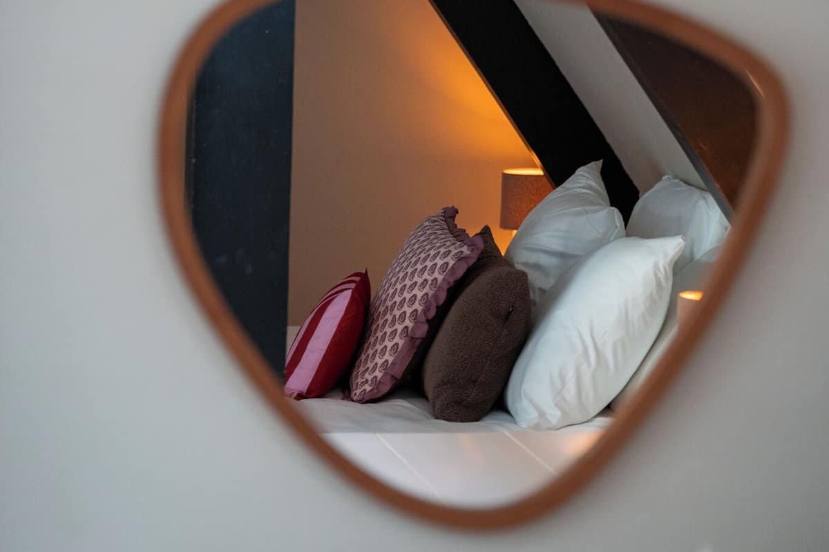 Espelho em formato de coração refletindo uma cama com almofadas e uma luminária ao fundo.