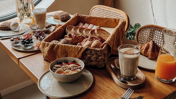 Een ontbijt opstelling met brood, gebak en drankjes op een houten tafel bij een raam.