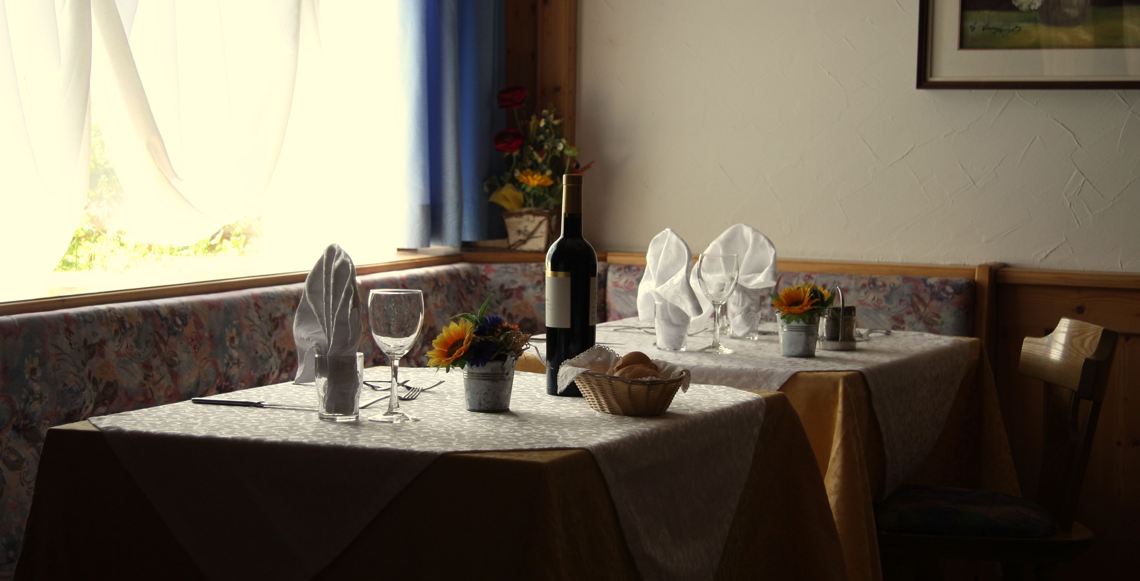 Une table à manger dressée avec une bouteille de vin, des verres et un panier de pain, avec une fenêtre et des rideaux en arrière-plan.