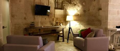 A cozy living room with stone walls, a flat-screen TV mounted on the wall, a wooden table with a lamp, and two sofas.