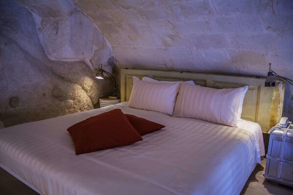 Una cama con ropa de cama blanca y dos almohadas, una lámpara de noche y una maleta sobre un suelo de madera.