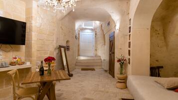 A narrow stone-walled hallway leading to a bedroom with a bed, a wooden table with a bottle and flowers, a TV mounted on the wall, and a chandelier.