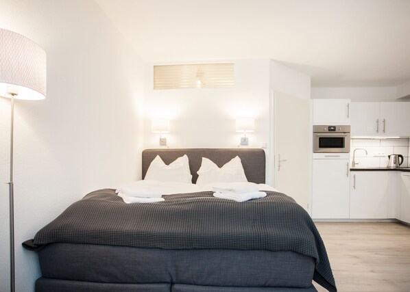 Une chambre d’hôtel comprenant un lit, des lampes de chevet, un luminaire fixé au mur, une fenêtre avec stores, une kitchenette équipée d’un four et d’un four à micro-ondes.