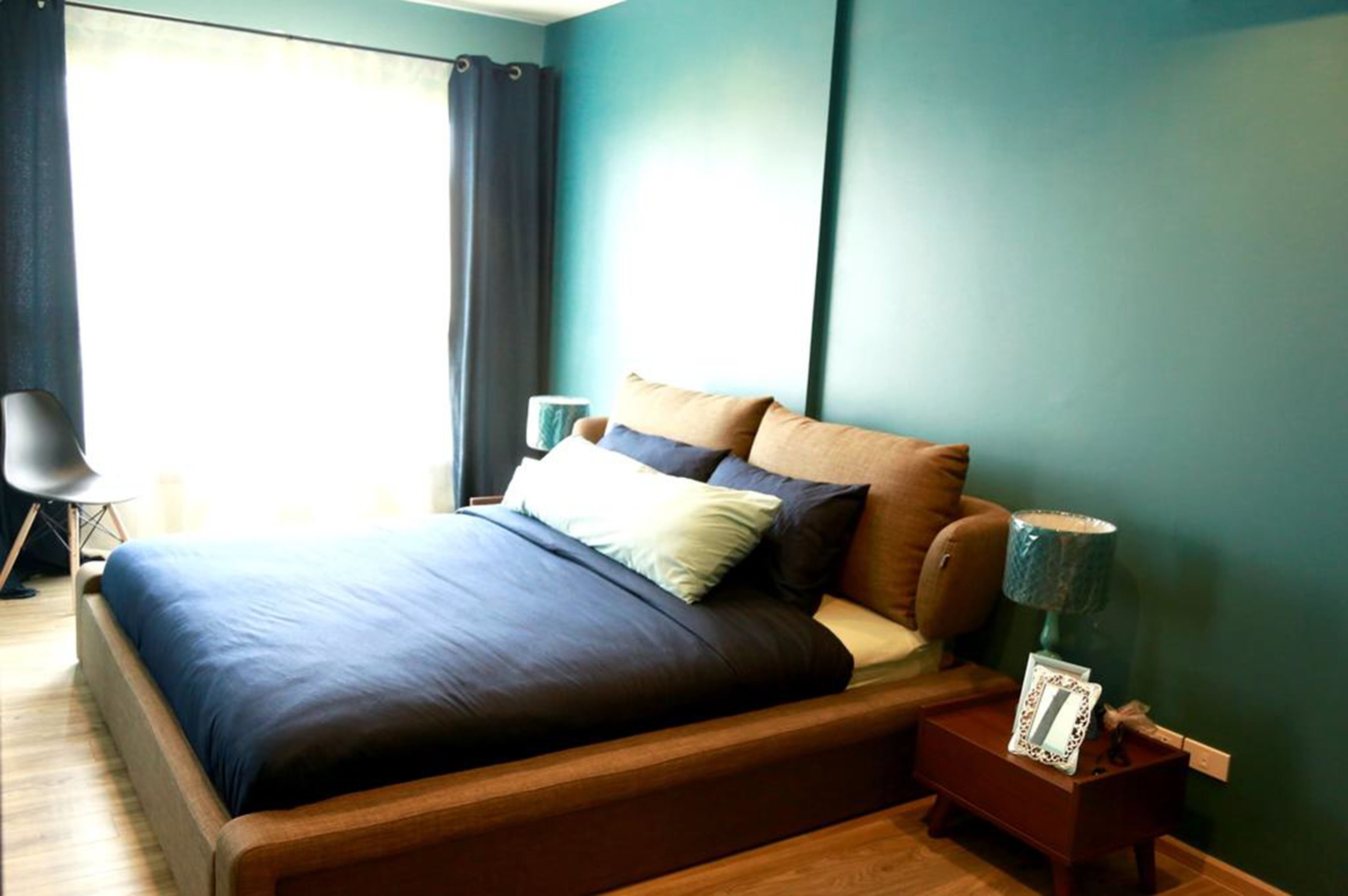 Cama bem arrumada com colcha azul escura e várias almofadas, mesa de cabeceira de madeira com abajur, e uma cadeira em um quarto com paredes verde-água e uma janela grande com cortinas.