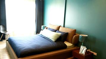 Cama bem arrumada com colcha azul escura e várias almofadas, mesa de cabeceira de madeira com abajur, e uma cadeira em um quarto com paredes verde-água e uma janela grande com cortinas.