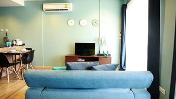 A living room with a blue sofa, a TV on a wooden stand, a dining table with chairs, and a wall-mounted air conditioner.
