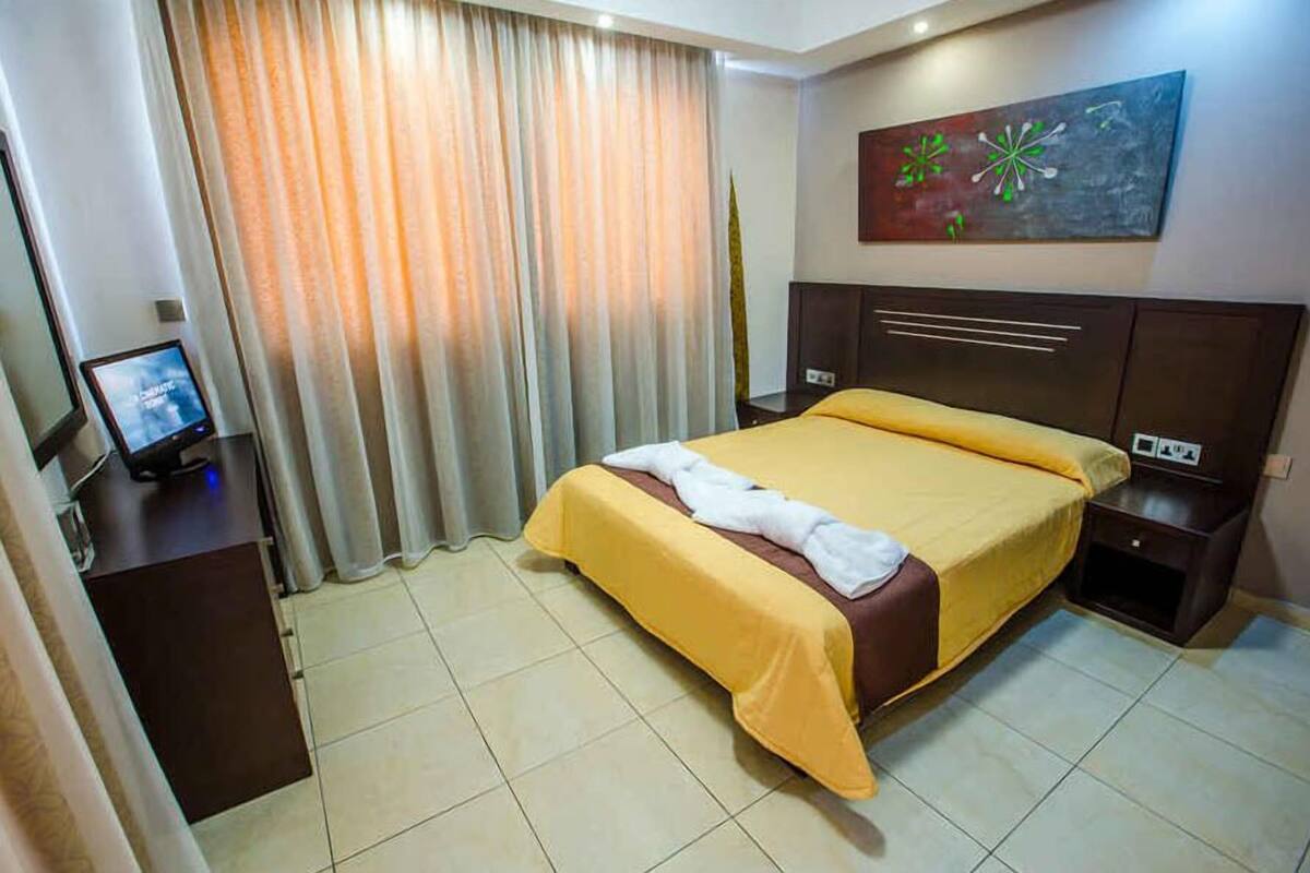 Una habitación de hotel con una cama, una mesita de noche con televisión, una lámpara, un cuadro en la pared y una ventana con cortinas.