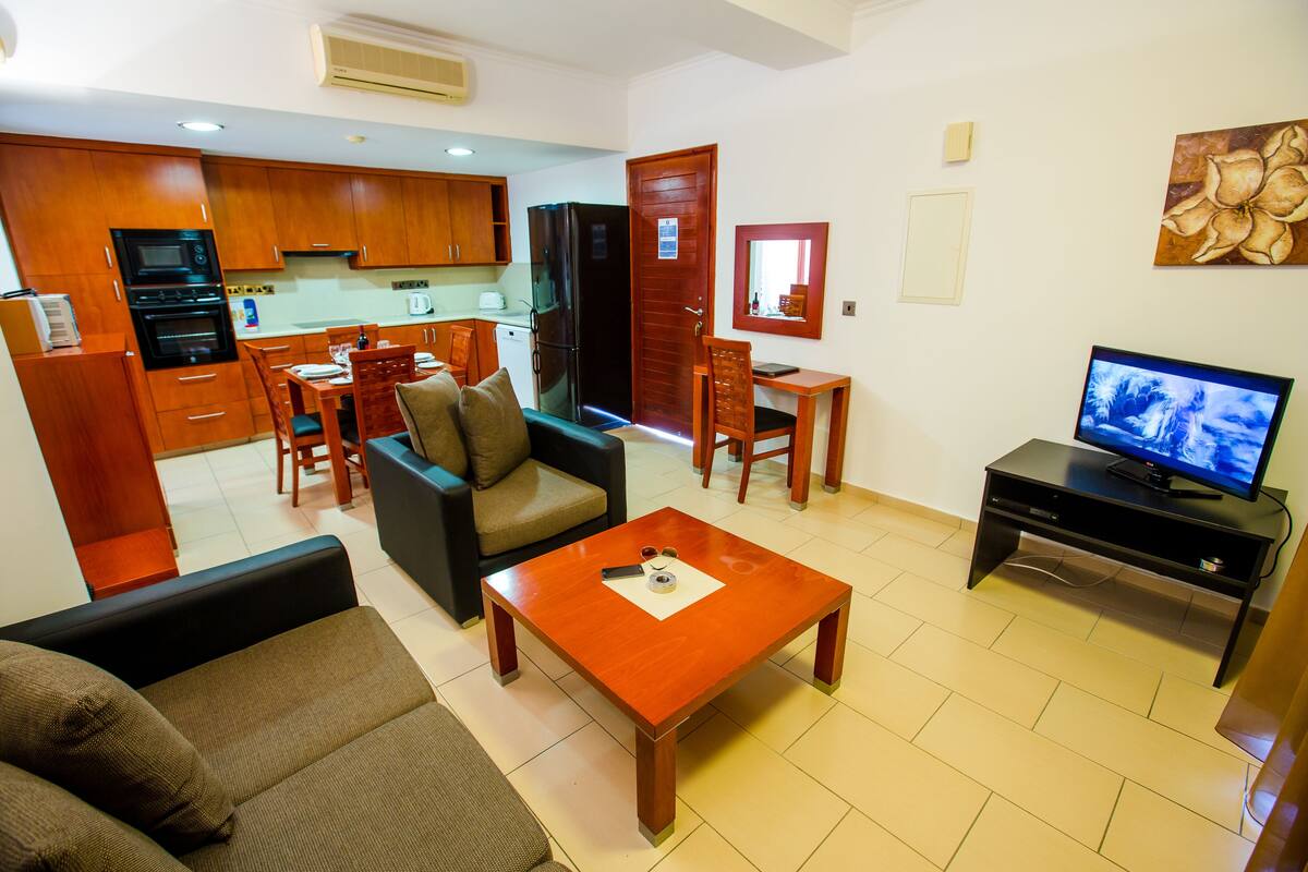Una sala de estar con sofá, mesa de centro, televisor y cocina al fondo.