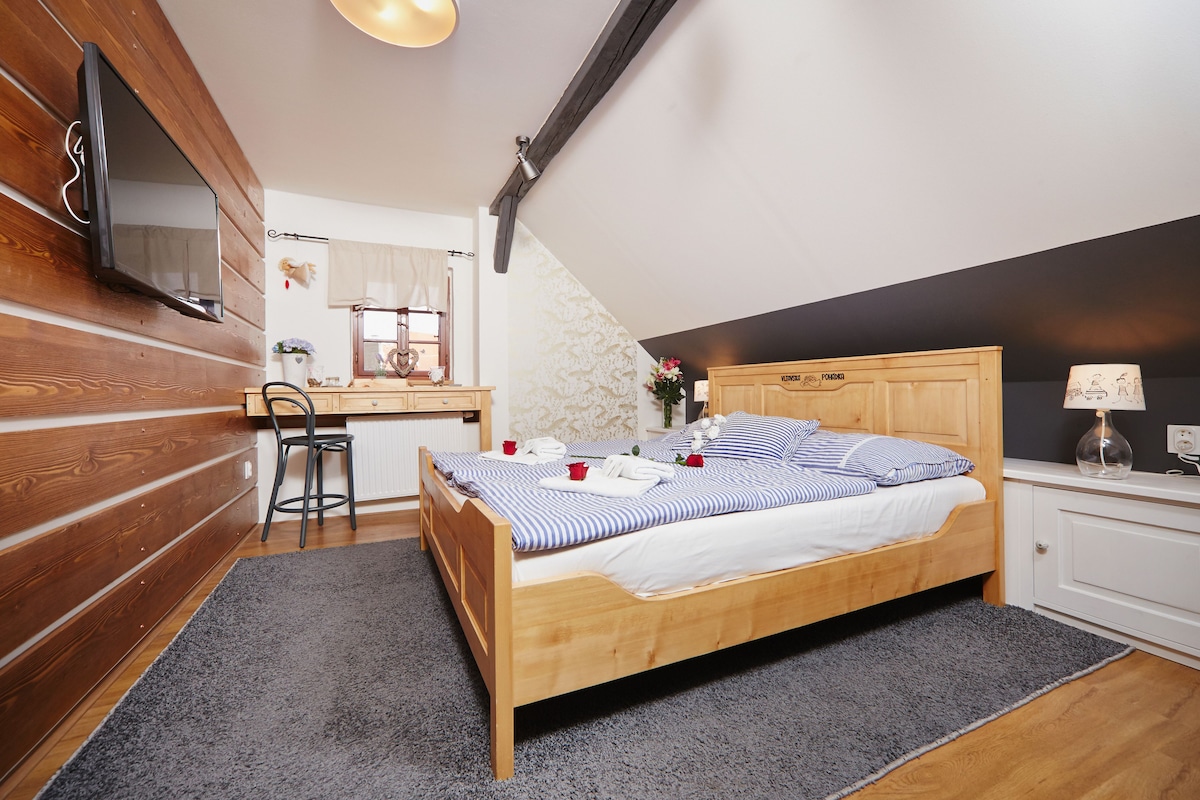 Un dormitorio con una cama de madera, un escritorio con silla, un televisor montado en la pared y una pequeña mesa con lámpara.