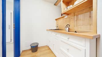 Cocina compacta con armarios blancos, encimera de madera, hervidor y cubo de basura.