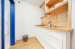 Cocina compacta con armarios blancos, encimera de madera, hervidor y cubo de basura.