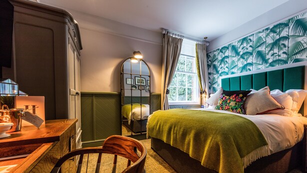 Ein Schlafzimmer mit Bett, Schreibtisch, Stuhl, einem Fenster mit Vorhängen und einer Wand mit Blättermuster.