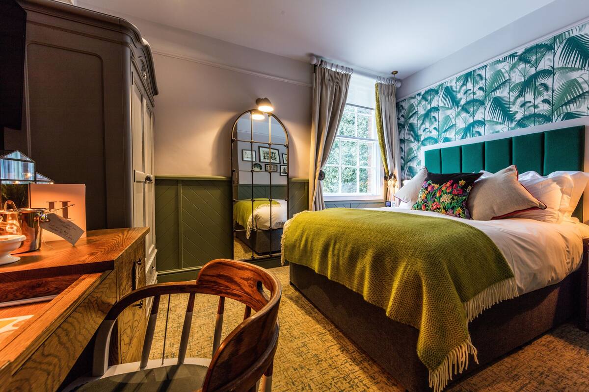 A bedroom with a bed, a desk, a chair, a window with curtains, and a wall with a leaf pattern.