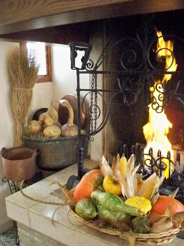 Una chimenea de piedra con fuego, una cesta de frutas y verduras, y un ambiente rústico con una escoba y una olla.