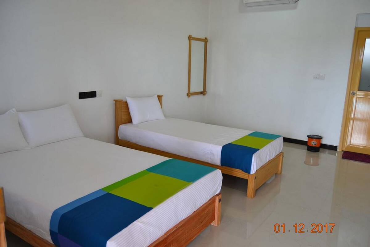 A hotel room with two single beds, each with a wooden frame and white bedding. There is a small round table with a red base and a black object on it. A wooden door is visible on the right side of the room.