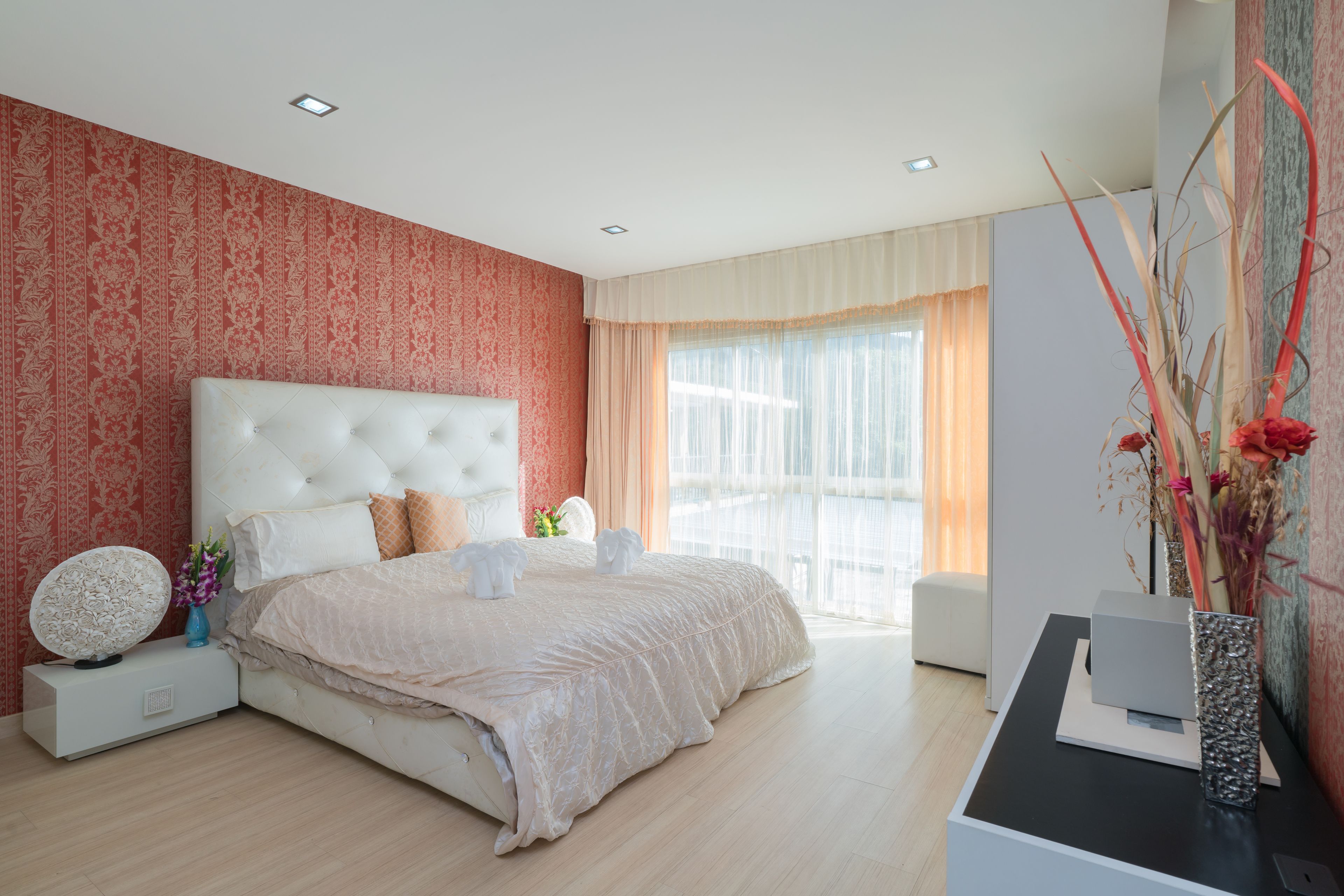 A modern bedroom with a large bed, a bedside table, a nightstand, a decorative vase, and a view of the outdoors through a window.