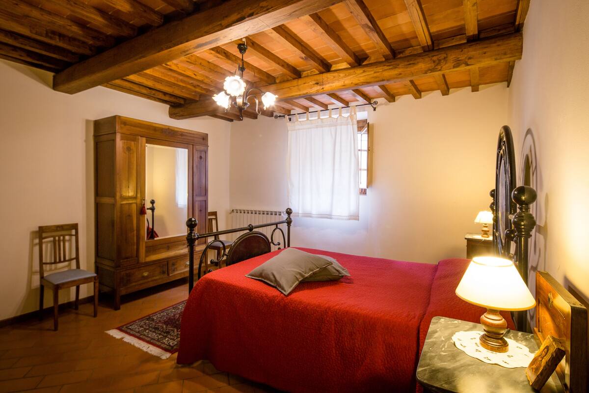 A bedroom with a wooden ceiling, a bed with a red bedspread, a nightstand with a lamp, a wooden wardrobe, and a chair.