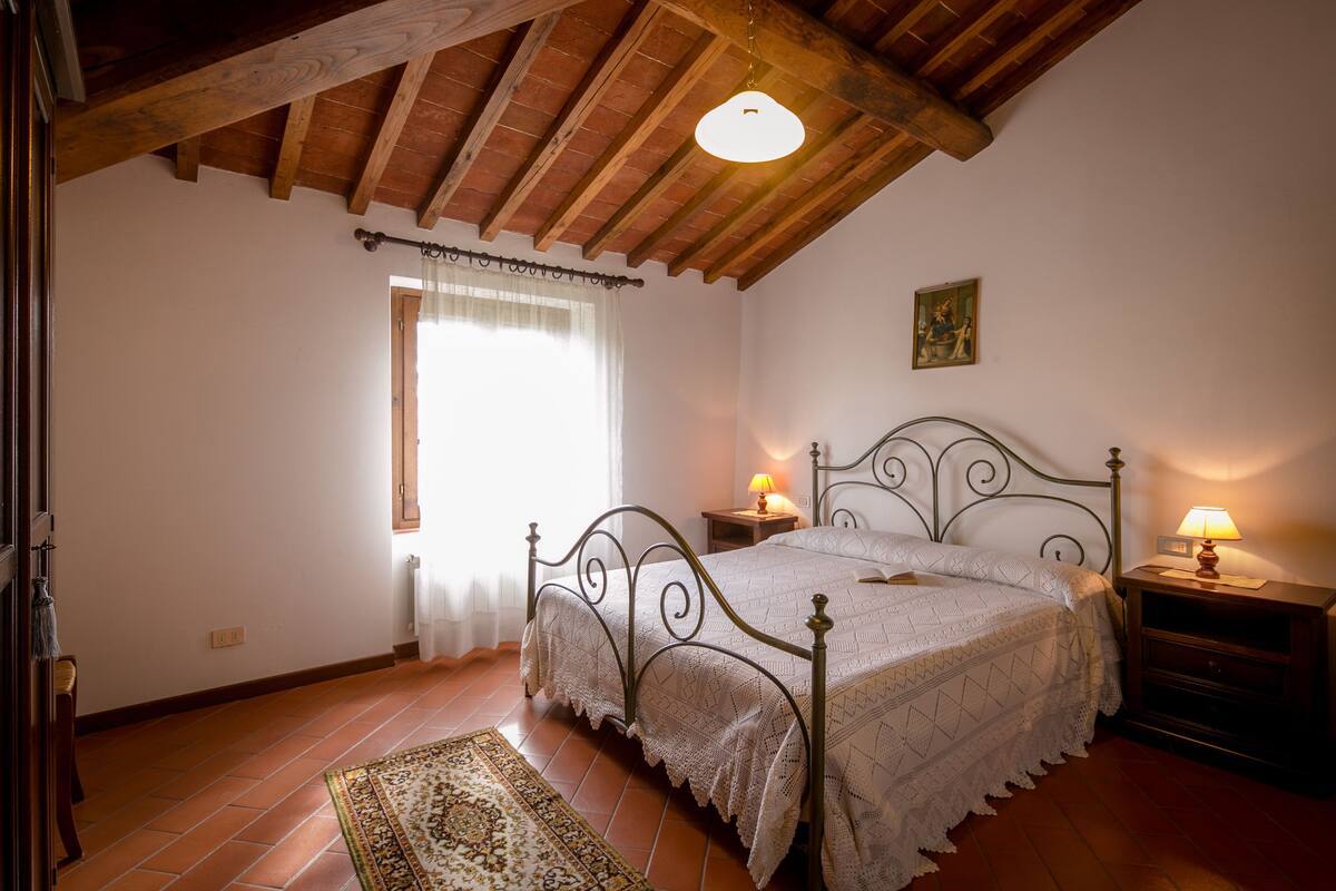 A bedroom with a wooden ceiling, a bed with a metal frame, a bedside table with a lamp, and a patterned rug on the floor.