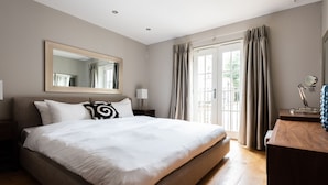 A bedroom with a large bed, a wooden dresser, a mirror, and a view of the outdoors through a window with curtains. - The Norfolk Townhouse - Large & Stunning 5BDR Mews Home on Private Street (London)