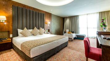 A hotel room with a large bed, a desk, a chair, a small table with fruit, and a vase with flowers.