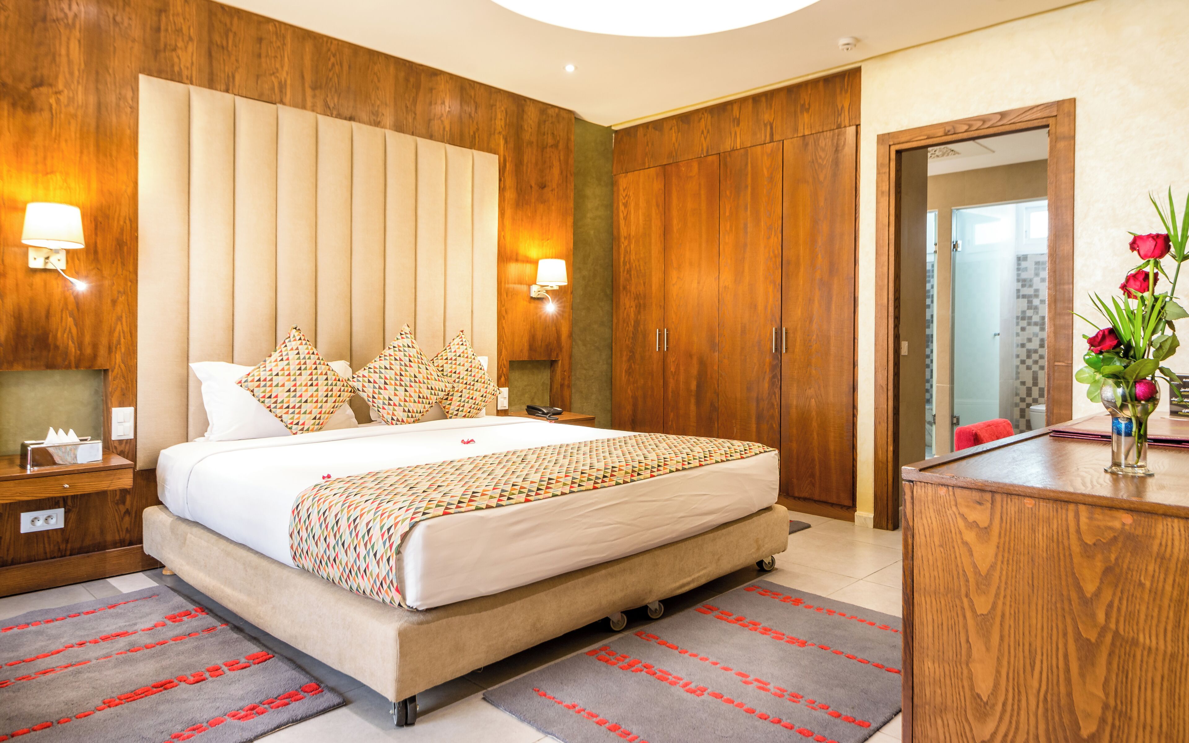A hotel room with a large bed, wooden wardrobe, bedside lamps, a mirror, and a vase with flowers.