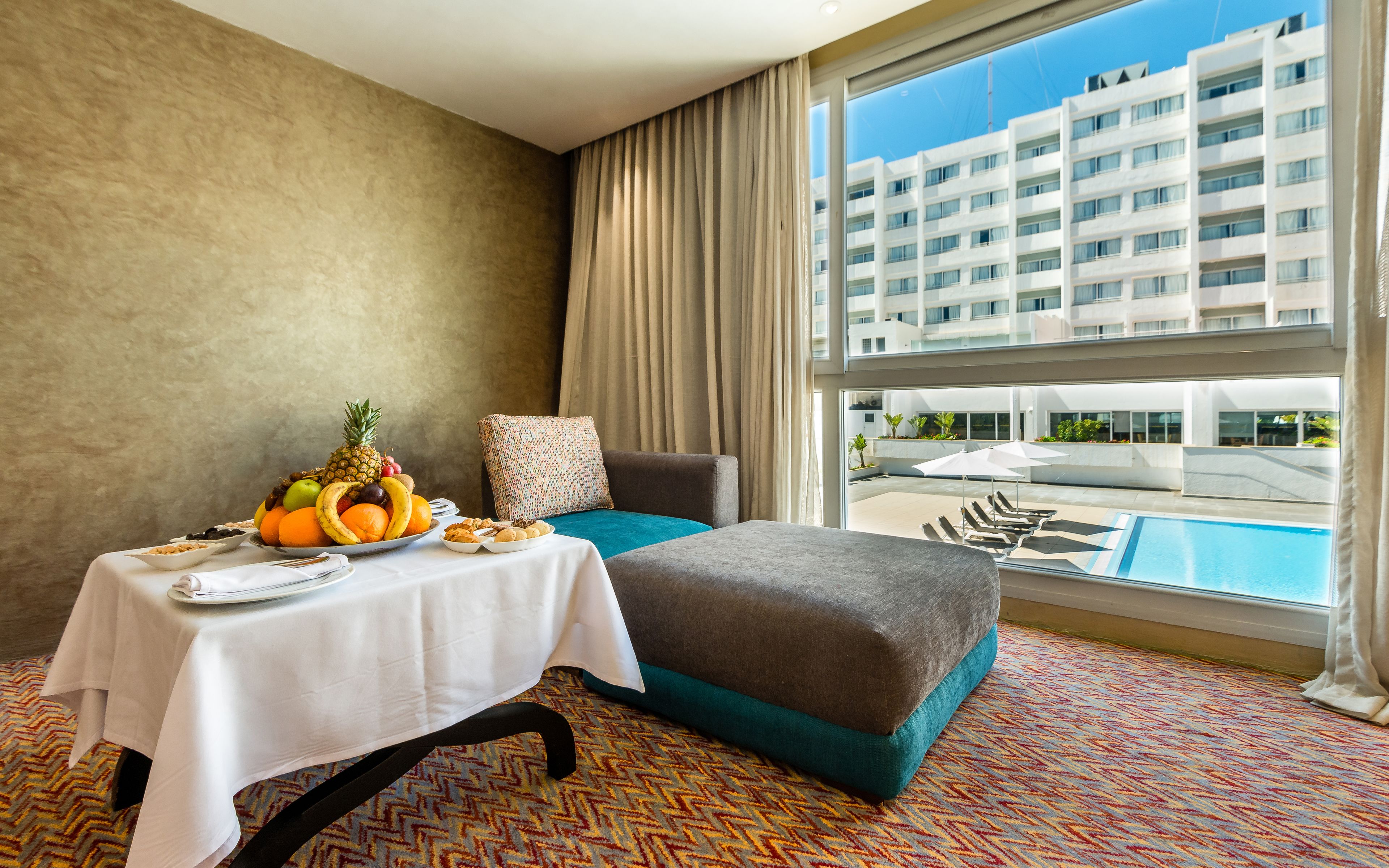 A hotel room with a pool view, a dining table with fruits and snacks, a sofa, and a patterned carpet.