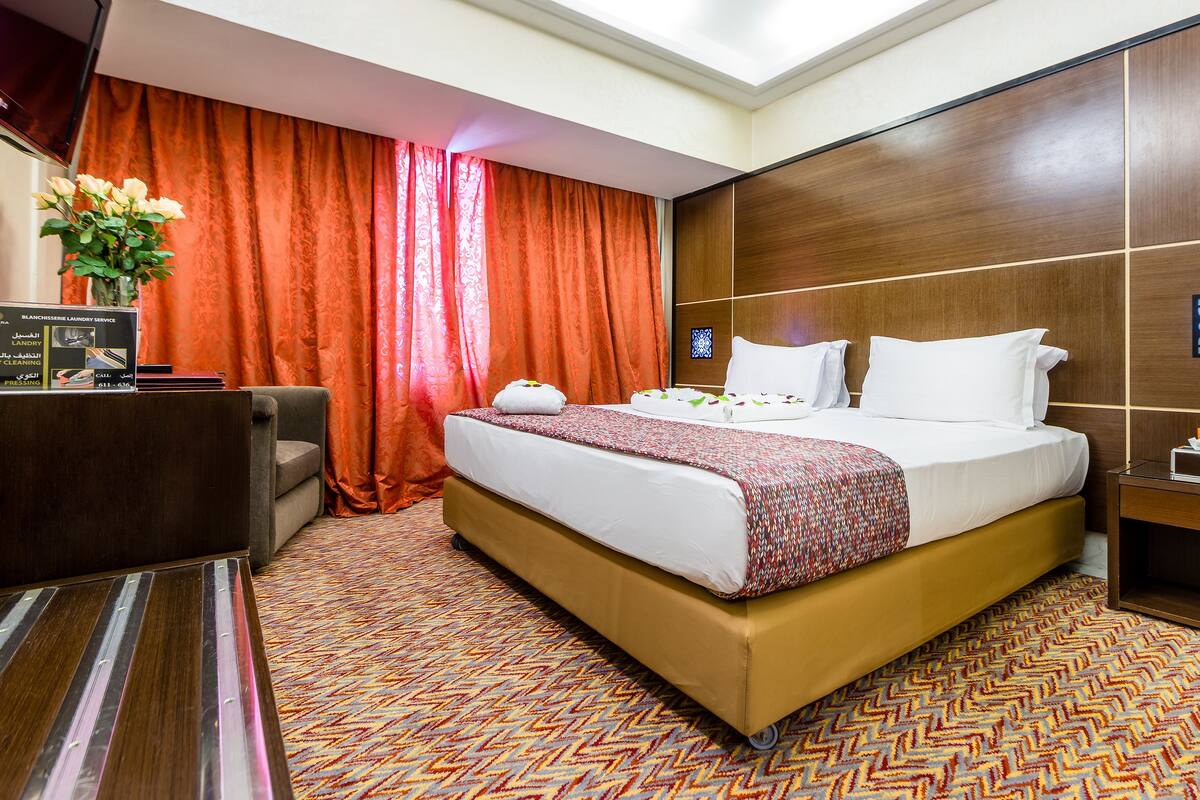 A hotel room with a bed, a desk, a chair, a nightstand, and a window with curtains.