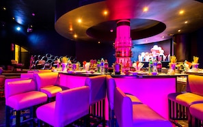 Uma área de bar com balcão central, diversas opções de assento e variedade de garrafas e copos.