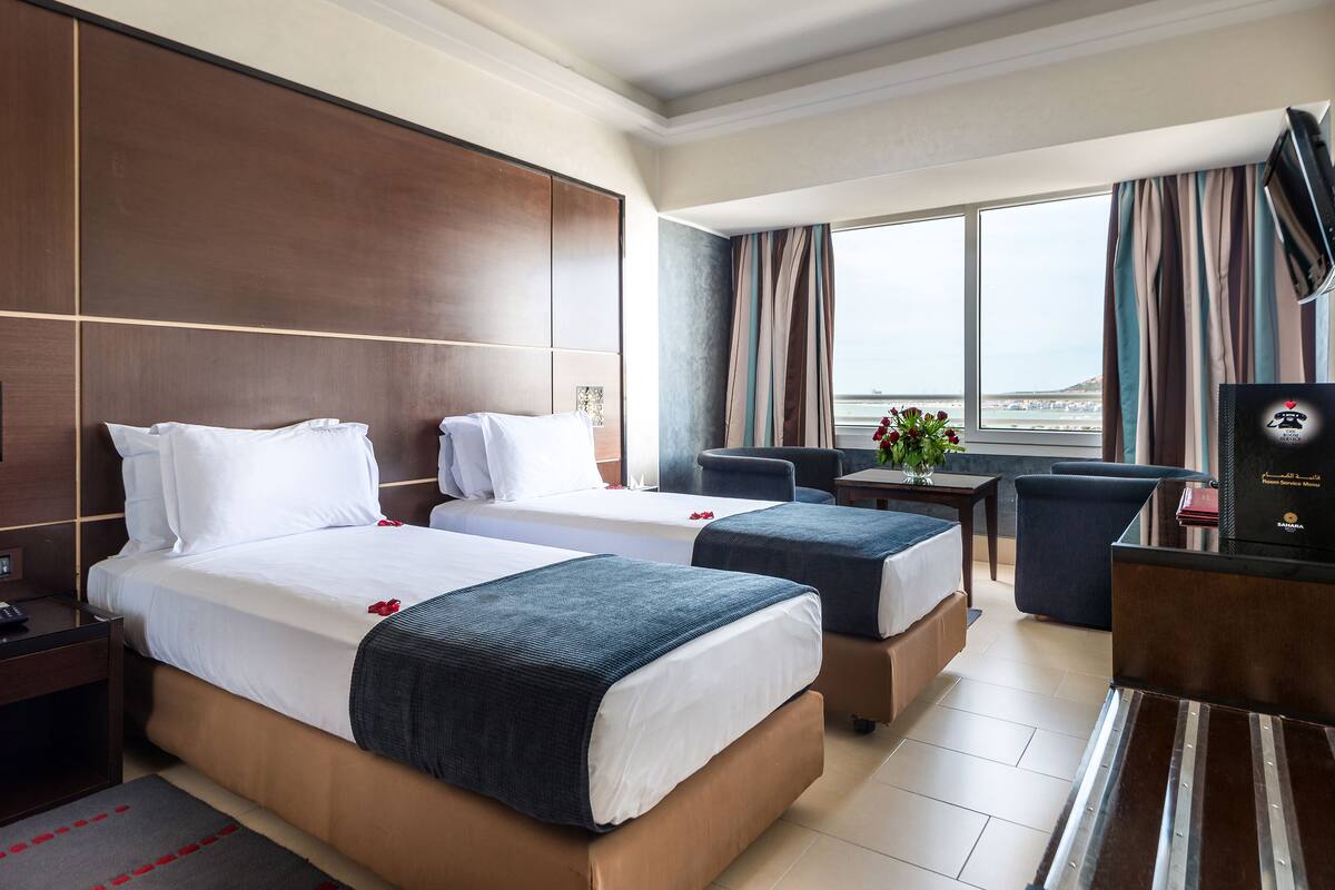 A hotel room with two beds, a desk, a chair, and a view of the sea.