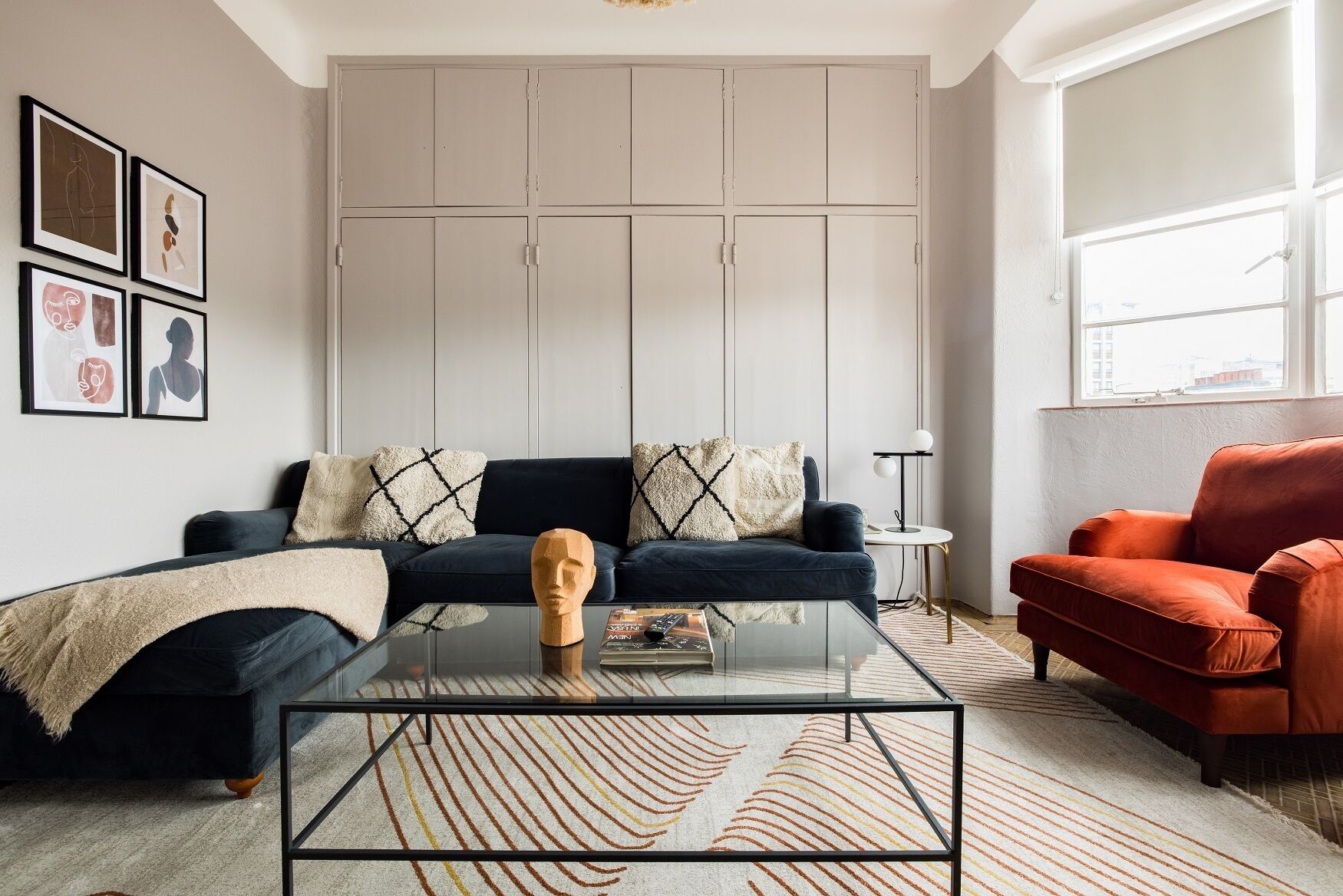 A modern living room with a dark blue sofa, a red armchair, a glass coffee table, and a large window providing natural light.